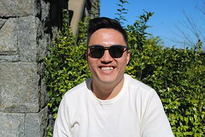 Trevor Wong wearing sunglasses and smiling with blue skies and a green bush in the background.